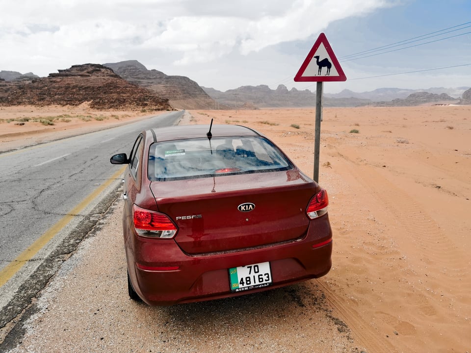 Eine rote Mietlimousine am sandigen Rand einer Wüstenstraße in Jordanien, neben einem Kamel-Warnschild, mit Felsbergen im Hintergrund.