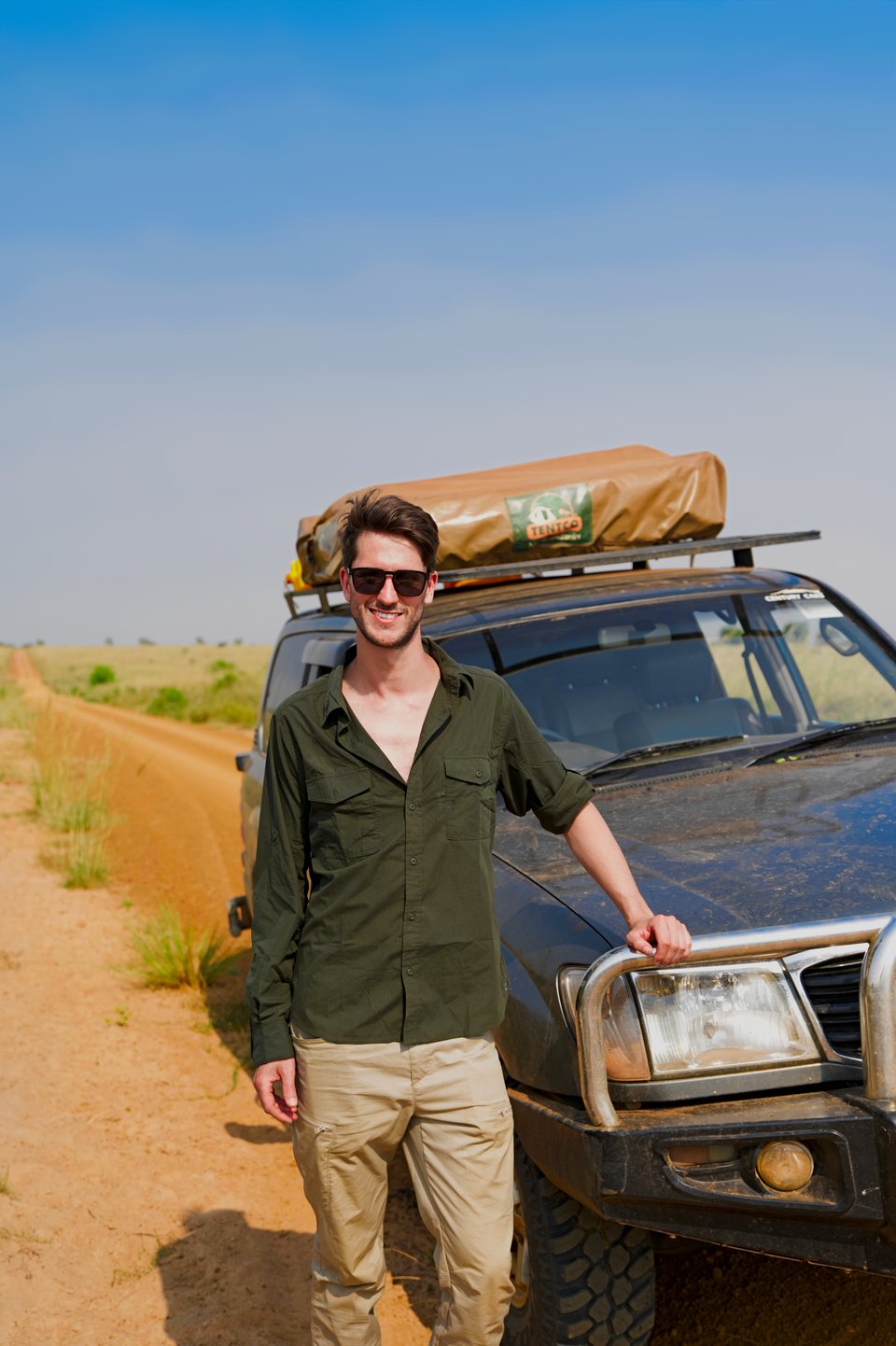 Martijn lehnt am Rammschutz eines Geländewagens mit Dachzelt, neben einer roten Piste durch das Grasland Ugandas.